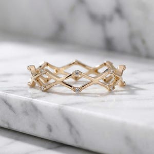 May include: A gold ring with a geometric design. The ring features diamond-shaped patterns and small, clear gemstones. The ring is displayed on a white marble surface.