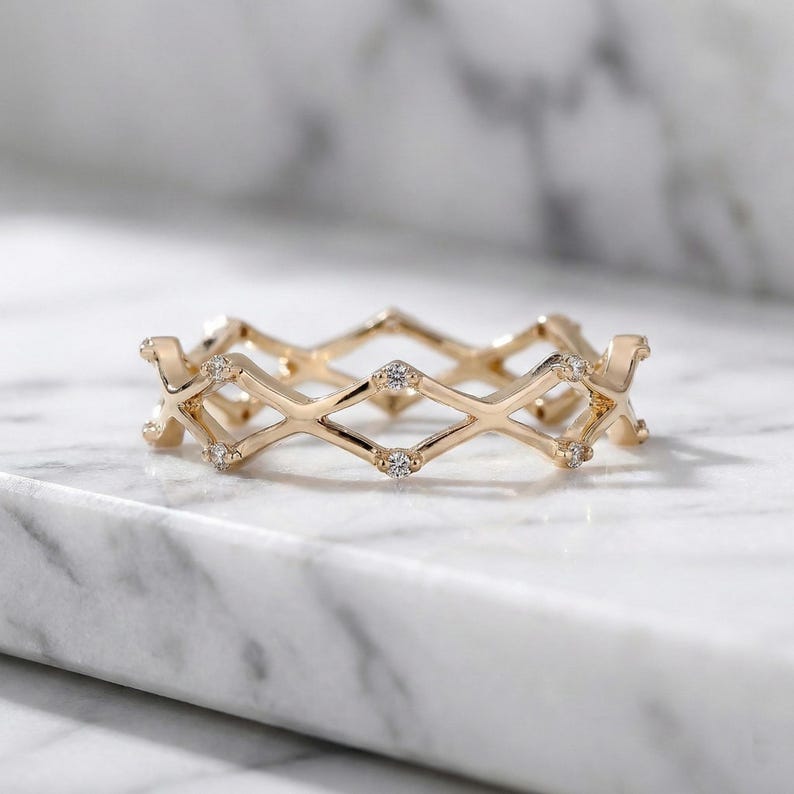 May include: A gold ring with a geometric design. The ring features diamond-shaped patterns and small, clear gemstones. The ring is displayed on a white marble surface.
