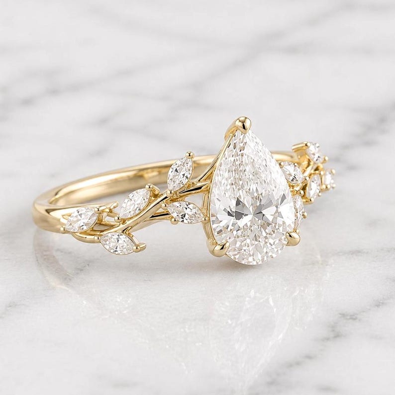 May include: A gold engagement ring featuring a large, pear-shaped diamond as the centerpiece. The band is adorned with smaller, marquise-cut diamonds and a delicate leaf design. The ring is set against a white marble background.