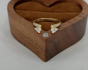 May include: A gold-colored ring with clear, sparkling stones and a unique open design. The ring is displayed inside a heart-shaped wooden jewelry box with a brown velvet interior. The ring features two pear-shaped stones.
