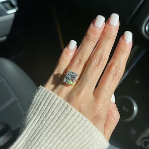 May include: A gold ring with a large, rectangular, clear gemstone. The ring is on a hand with white nail polish, and the person is wearing a cream-colored sweater. The ring is catching the sunlight.