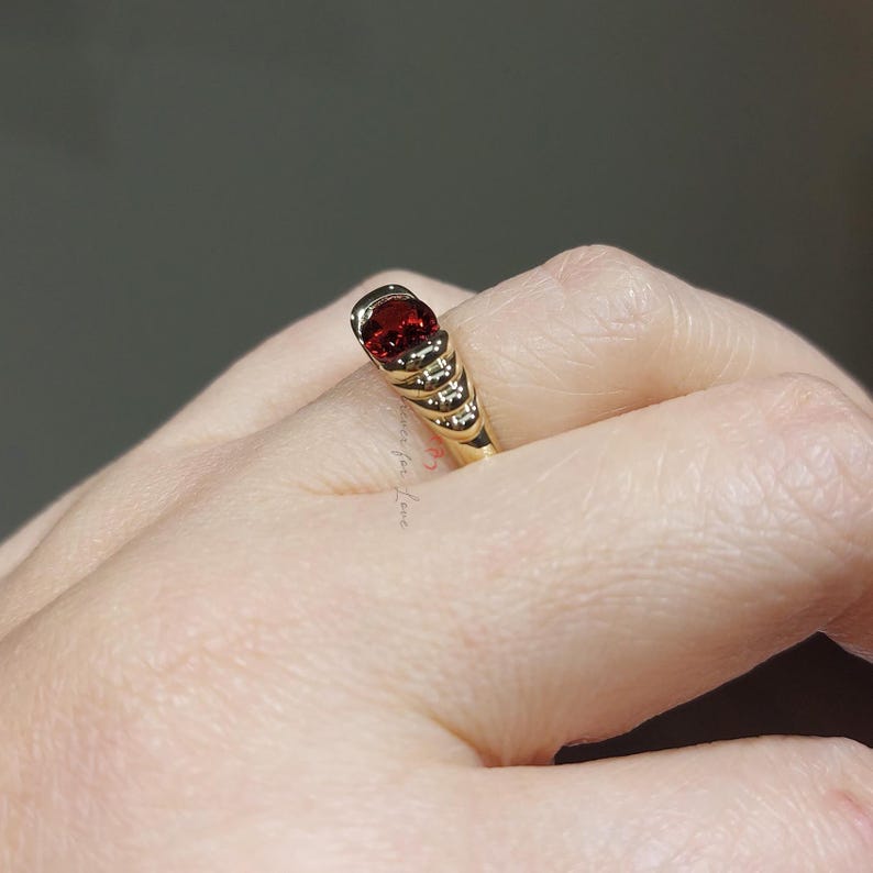 Puede incluir: Un anillo de oro con una gran piedra preciosa roja de corte ovalado. La banda del anillo tiene un dise&ntilde;o texturizado con peque&ntilde;as piedras de acento transparentes. El anillo se muestra en un dedo, destacando su elegante dise&ntilde;o y color.