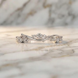 Può includere: Un delicato anello d'argento con un design ispirato alla natura. L'anello presenta una serie di rami intrecciati adornati con piccole gemme scintillanti. Le gemme sono raggruppate in vari punti lungo la fascia dell'anello.
