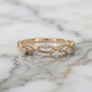 May include: A gold ring with a detailed vine pattern. The ring has three small, clear, round gemstones. The ring is shown on a marble surface, reflecting light. The ring's design is reminiscent of nature.