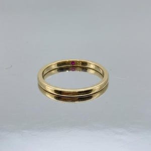 May include: A gold band ring with a small red gemstone. The ring is simple and elegant, with a polished finish. The ring is shown on a reflective surface.