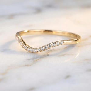 May include: A curved gold ring with a row of small, clear gemstones set along the top curve. The ring has a smooth, polished finish and is set against a blurred, light-colored background. The ring's design is elegant and minimalist.