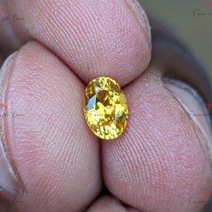 May include: A close-up of a yellow oval gemstone with facets. The gemstone is held between two fingers.