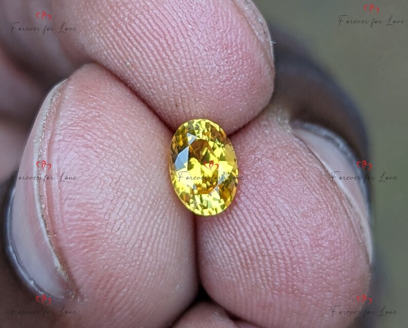 May include: A close-up of a yellow oval gemstone with facets. The gemstone is held between two fingers.