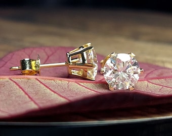 May include: A pair of gold stud earrings with a clear round gemstone in each. The earrings are on a pink leaf.