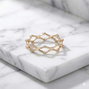 May include: A gold ring with a geometric design. The ring features diamond-shaped cutouts and small, clear gemstones. The ring is displayed on a white marble surface, highlighting its intricate design and elegant style.