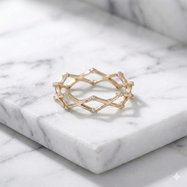 May include: A gold ring with a geometric design. The ring features diamond-shaped cutouts and small, clear gemstones. The ring is displayed on a white marble surface, highlighting its intricate design and elegant style.