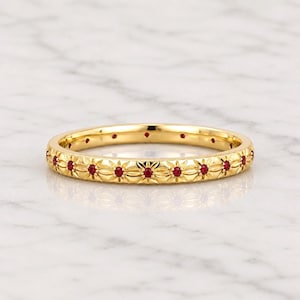 May include: A gold band ring featuring a repeating floral pattern engraved along the exterior. Small, round, vibrant red rubies are set into the centre of each engraved flower, creating a continuous decorative motif around the entire circumference.