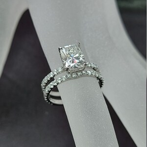 May include: A radiant-cut diamond engagement ring with a pavé band, paired with a matching diamond band. The rings are set in a silver metal, showcasing the brilliance of the clear gemstones. The design is elegant and classic.