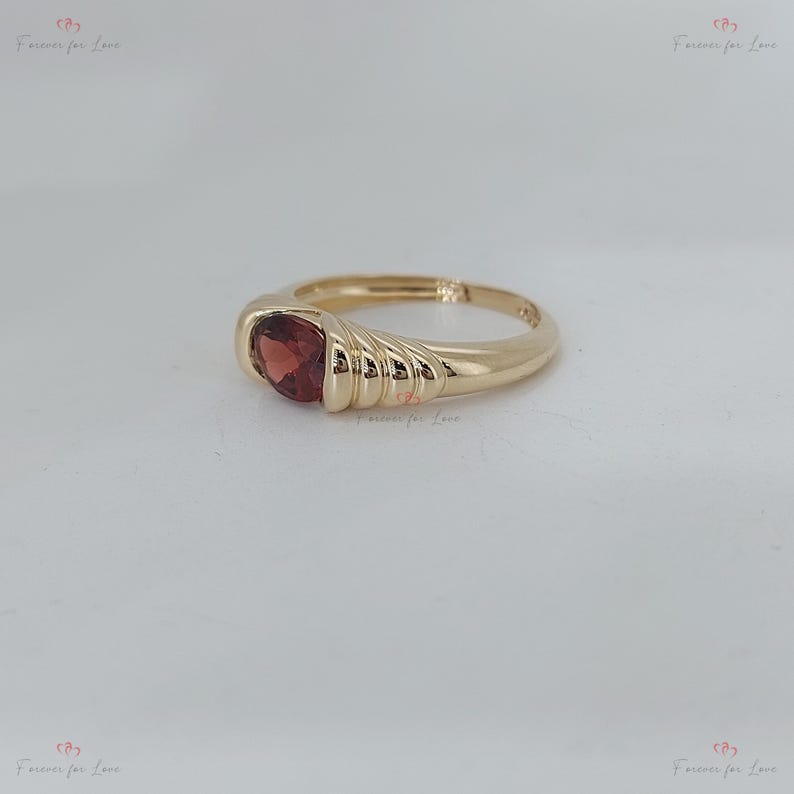 Puede incluir: Un anillo de oro con una piedra preciosa roja oscura en forma de coraz&oacute;n. El anillo tiene una banda lisa y pulida que se ensancha para sostener la piedra. El engaste presenta un dise&ntilde;o acanalado en el lateral, agregando textura. El anillo est&aacute; sobre una superficie blanca.