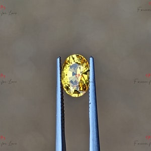 May include: A yellow oval gemstone held by tweezers. The gemstone has a slight golden hue.