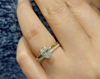 May include: A gold engagement ring with an oval-cut diamond center stone and a diamond-studded band. The ring is on a finger, with a matching gold band. The background is a blue textured fabric.