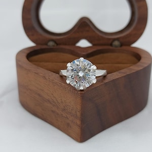 Puede incluir: Un anillo de compromiso plateado con una gema grande, redonda y transparente. El anillo se presenta en un joyero de madera en forma de corazón. La caja está abierta, revelando el anillo. El fondo es claro.