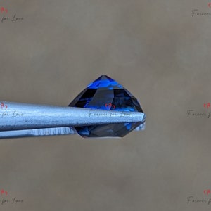 May include: A deep blue gemstone with a faceted cut, held by tweezers. The gemstone is a dark blue color with a slight purple hue.