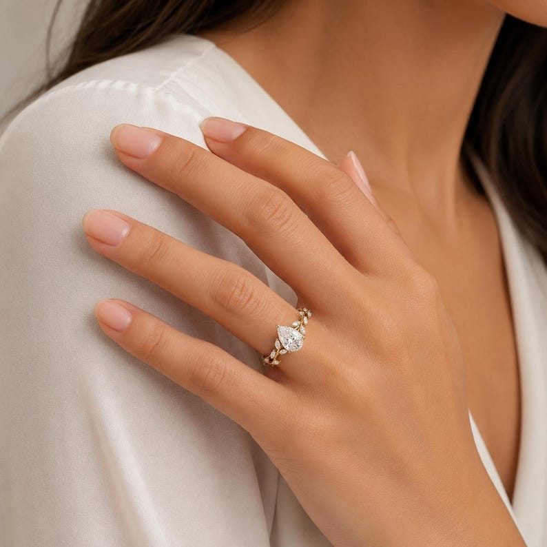 May include: A gold ring with a pear-shaped diamond center stone. The band features a delicate leaf design with smaller accent diamonds. The ring is worn on a person's finger, showcasing its elegant and intricate details.