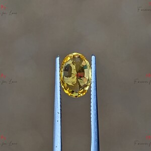 May include: A yellow oval gemstone held by tweezers. The gemstone is transparent and has a slight sparkle.