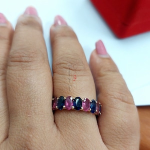 Puede incluir: Un anillo de banda de oro con piedras ovaladas azules y rosadas alternadas. El anillo está en el dedo de una persona.