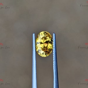 May include: A yellow oval gemstone held by tweezers. The gemstone has a faceted cut and a bright, golden hue.