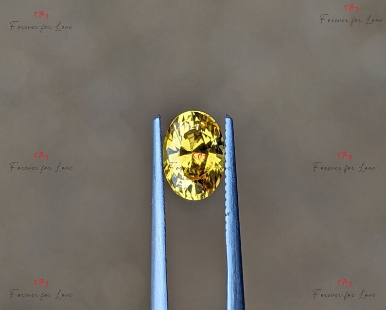 May include: A yellow oval gemstone held by tweezers. The gemstone has a faceted cut and a bright, golden hue.