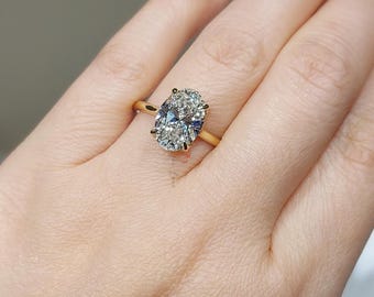 May include: A gold ring featuring a large, oval-cut diamond. The diamond is clear and sparkling, set on a thin, yellow gold band. The ring is shown on a person's finger, with a neutral background.
