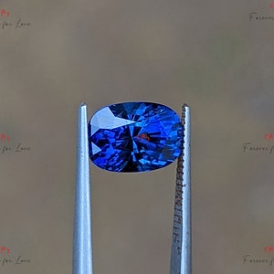 May include: A deep blue oval sapphire gemstone held in tweezers. The sapphire has a faceted cut and a brilliant shine.
