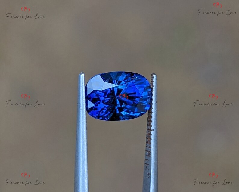 May include: A deep blue oval sapphire gemstone held in tweezers. The sapphire has a faceted cut and a brilliant shine.