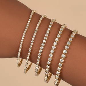 Puede incluir: Cinco pulseras doradas con piedras preciosas redondas y transparentes. Las pulseras var&iacute;an en tama&ntilde;o, con las piedras preciosas muy juntas. Las pulseras se muestran sobre un tono de piel marr&oacute;n, resaltando el brillo y el dise&ntilde;o.