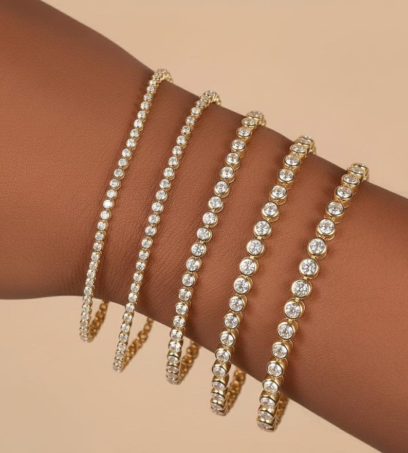 Puede incluir: Cinco pulseras doradas con piedras preciosas redondas y transparentes. Las pulseras var&iacute;an en tama&ntilde;o, con las piedras preciosas muy juntas. Las pulseras se muestran sobre un tono de piel marr&oacute;n, resaltando el brillo y el dise&ntilde;o.