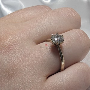 May include: A gold engagement ring with a round, clear diamond center stone. The ring is on a finger.