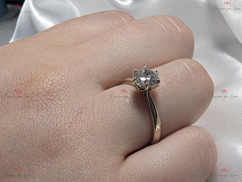 May include: A gold engagement ring with a round, clear diamond center stone. The ring is on a finger.