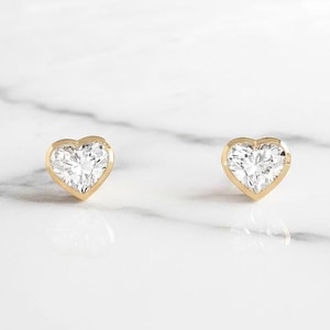 May include: A pair of heart-shaped stud earrings. Each earring has a clear, faceted gemstone set in a gold bezel. The earrings are displayed on a white marble surface, creating a simple and elegant presentation.