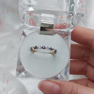 May include: A gold band ring with a row of alternating blue and white gemstones. The ring is displayed in a white ring box.