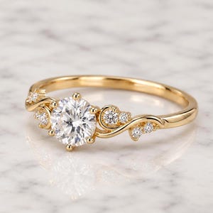 May include: A gold ring featuring a large round brilliant-cut center stone flanked by smaller round accent stones set along a delicate, swirling band. The metal has a polished finish, and the design incorporates multiple prongs to secure the stones.