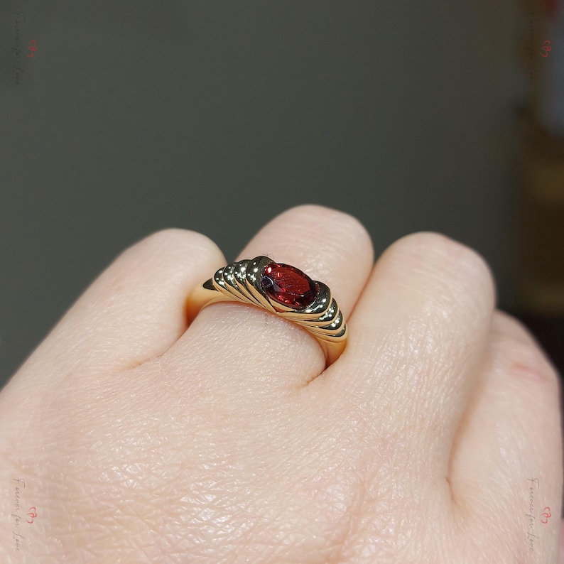 Puede incluir: Un anillo de oro con una llamativa gema roja ovalada. La banda del anillo tiene un dise&ntilde;o texturizado y acanalado que enmarca la piedra central. El anillo se muestra en un dedo, destacando su estilo elegante y cl&aacute;sico.