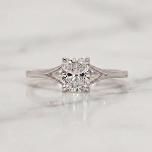 Op de afbeelding: Een verlovingsring met een rechthoekig geslepen diamant. De ring heeft een split-shank band van glanzend, lichtgekleurd metaal, waarschijnlijk platina of witgoud. De diamant is het middelpunt en reflecteert het licht. Een prachtig sieraad.