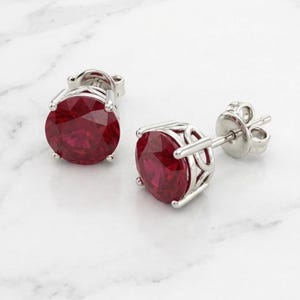 Peut inclure: Une paire de boucles d'oreilles à clous avec des pierres rondes à facettes d'un rouge profond, serties dans des paniers en métal argenté poli. Chaque boucle possède un fermoir à tige et une galerie ajourée décorative maintenant la pierre.