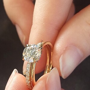 May include: A gold engagement ring with a large, round, clear diamond center stone and a smaller band of diamonds set in a pave setting. The ring is worn on a finger with a gold wedding band.