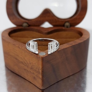 Op de afbeelding: Een zilveren ring met twee smaragdgeslepen diamanten en een band bezet met kleinere diamanten. De ring wordt gepresenteerd in een hartvormige houten sieradendoos.
