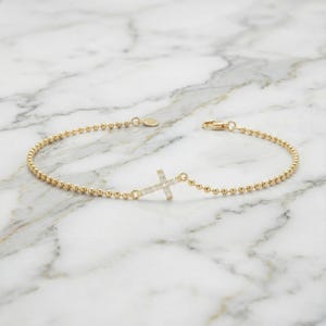 Op de afbeelding: Een gouden armband met kralenketting en een centrale kruishanger ingelegd met kleine heldere steentjes. De armband bestaat uit een fijne gouden ketting van kleine bolvormige kraaltjes en een veilige sluiting met een karabijnhaak.