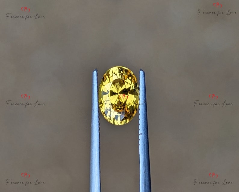 May include: A yellow oval gemstone held by tweezers. The gemstone has a faceted cut and a bright, golden color.
