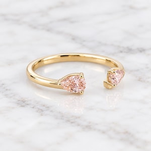 Puede incluir: Un anillo abierto de oro con dos piedras preciosas de color melocotón en talla pera situadas en cada extremo de la banda. El anillo tiene un acabado en oro amarillo pulido y las piedras están aseguradas con pequeñas garras.
