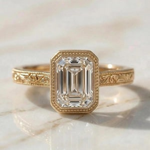 May include: A gold ring featuring an emerald-cut diamond. The ring has an engraved band and a bezel setting. The clear, rectangular diamond is the focal point, set against a marble surface. The ring's design is intricate.