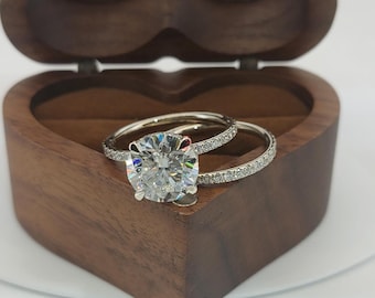 Könnte beinhalten: Nahaufnahme eines Diamant-Verlobungsringes und eines Eherings in einer herzförmigen Holzschachtel. Der Verlobungsring hat einen großen, runden Brillanten, der von einem Halo aus kleineren Diamanten umgeben ist. Der Ehering ist ein schlichtes, dünnes Band mit einer Reihe kleiner Diamanten.