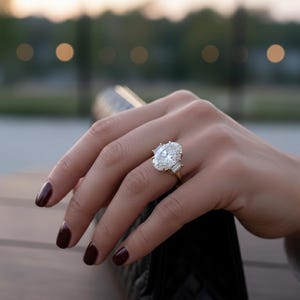 May include: A gold ring featuring a large oval-cut diamond center stone, flanked by two smaller baguette-cut diamonds. The ring is set on a gold band and is displayed on a hand with dark red nail polish.