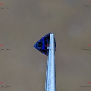 May include: A close-up of a deep blue gemstone held in tweezers. The gemstone is faceted and has a pointed shape.