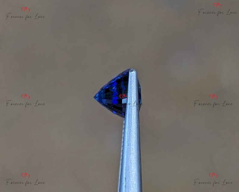 May include: A close-up of a deep blue gemstone held in tweezers. The gemstone is faceted and has a pointed shape.